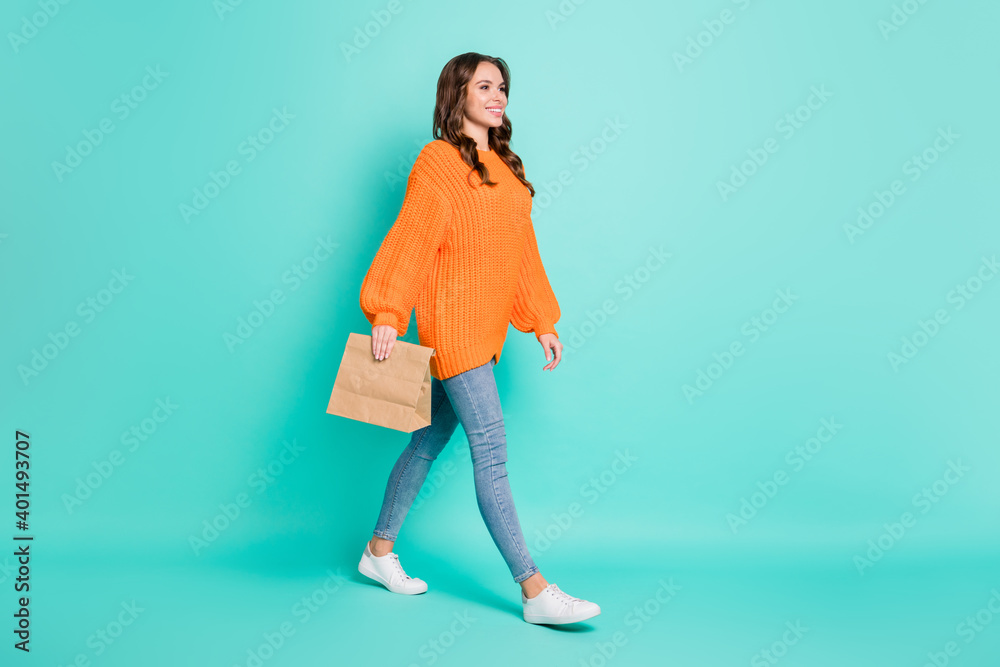 Full size profile side photo of young lovely pretty smiling girl go walk with paper bag in hand isolated on teal color background Full size profile side photo of young lovely pretty smiling girl go walk with paper bag in hand isolated on teal color background