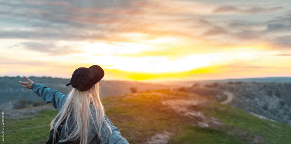 Back view of young happy blonde girl with black cap, on peak of hills at sunset. Travel concept. Panoramic photo with copy space.