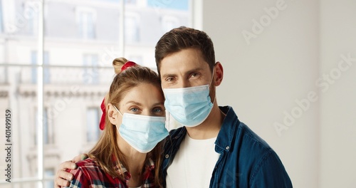 Close up portrait of happy Caucasian young married couple man and woman in medical masks standing in new apartment hugging and looking at camera. Renovation concept. Redesigning room. Home repair