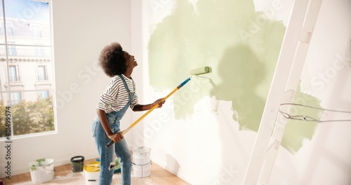 Young African American woman dancing and painting wall with roller brush while renovating apartment. Rear of female having fun redecorating home, renovating and improving Repair and decorating concept photo