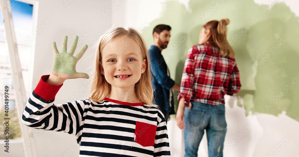 Close up portrait of joyful little cute girl smiling to camera showing hand palm in green paint. Mom and dad painting wall on background. Home repair and remodeling. Renewing house. Interior design Close up portrait of joyful little cute girl smiling to camera showing hand palm in green paint. Mom and dad painting wall on background. Home repair and remodeling. Renewing house. Interior design