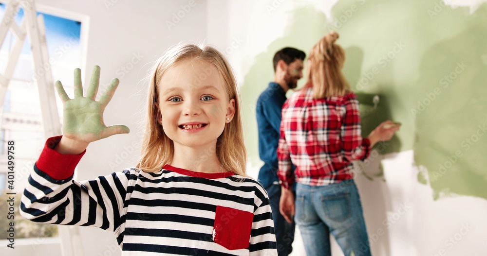 Close up portrait of joyful little cute girl smiling to camera showing hand palm in green paint. Mom and dad painting wall on background. Home repair and remodeling. Renewing house. Interior design Close up portrait of joyful little cute girl smiling to camera showing hand palm in green paint. Mom and dad painting wall on background. Home repair and remodeling. Renewing house. Interior design