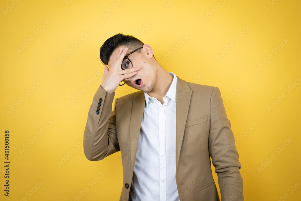 Young business man over isolated yellow background peeking in shock covering face and eyes with hand, looking through fingers with embarrassed expression Young business man over isolated yellow background peeking in shock covering face and eyes with hand, looking through fingers with embarrassed expression