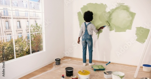 Young African American female alone painting wall with roller brush in olive color while renovating apartment. Rear of woman redecorating home, renovating and improving Repair and decorating concept