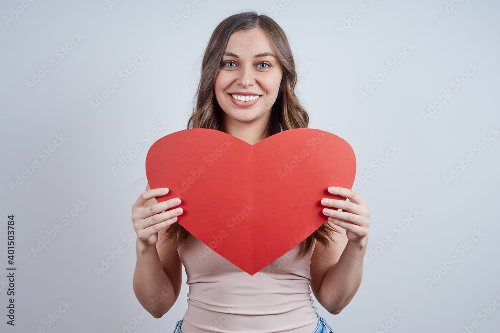 Smiling blonde woman holding a paper heart at her chest, looking at the camera, on gray background. Happy Valentine's Day. World heart day. Smiling blonde woman holding a paper heart at her chest, looking at the camera, on gray background. Happy Valentine's Day. World heart day.