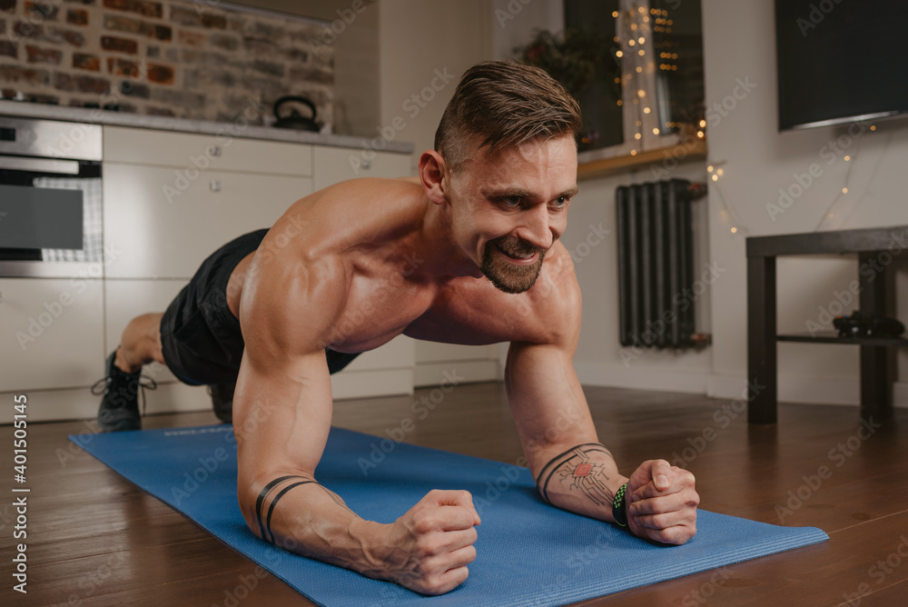 A happy muscular man with a naked torso is doing a plank on a blue yoga mat in his apartment in the evening. A smiling bodybuilder with tattoos on his forearms is training at home. A happy muscular man with a naked torso is doing a plank on a blue yoga mat in his apartment in the evening. A smiling bodybuilder with tattoos on his forearms is training at home.