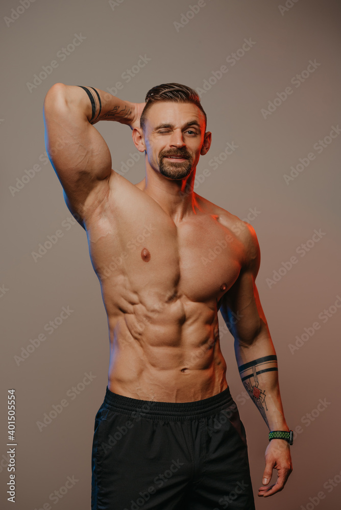 A smiling muscular man with a beard is posing. The athletic guy is demonstrating his sporty physique. A bodybuilder with tattoos on his forearms is standing with one hand behind his head