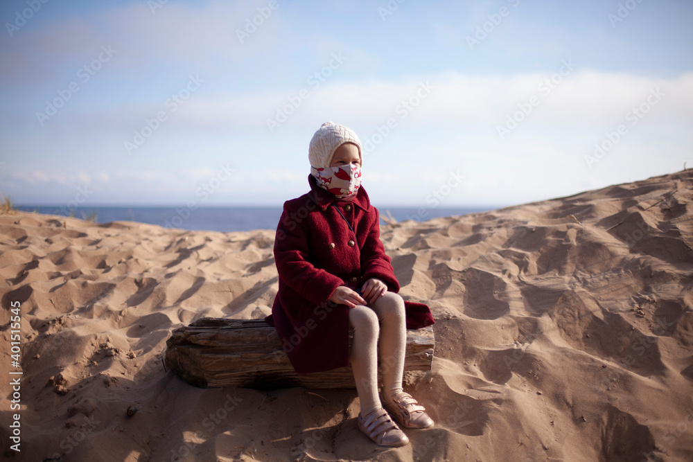 Baby girl in a funny face mask sitting near the sea on the beach. Self-isolation. Outdoor fun during coronavirus outbreak. New normal and social distancing in covid-19. 