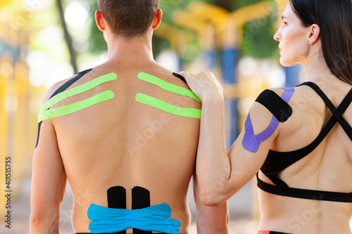 Back view of brunette woman holding hand on shoulder of unrecognizable man, professional caucasian athletes with colorful kinesiological taping on bodies, posing at sports ground, summer day. photo