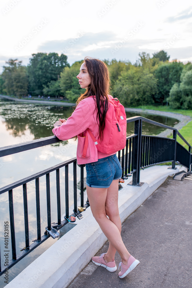 woman in summer in city near lake, river pond, looks at landscape, pink backpack behind her back. Relaxation after work, enjoying nature.