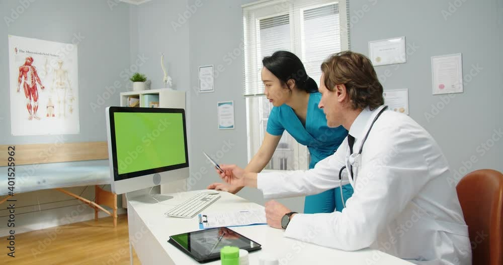 Side view of Caucasian man healthcare specialist doctor sitting in cabinet in medical center and looking at computer green screen, talking to Asian female assistant about patient diagnosis, chroma key