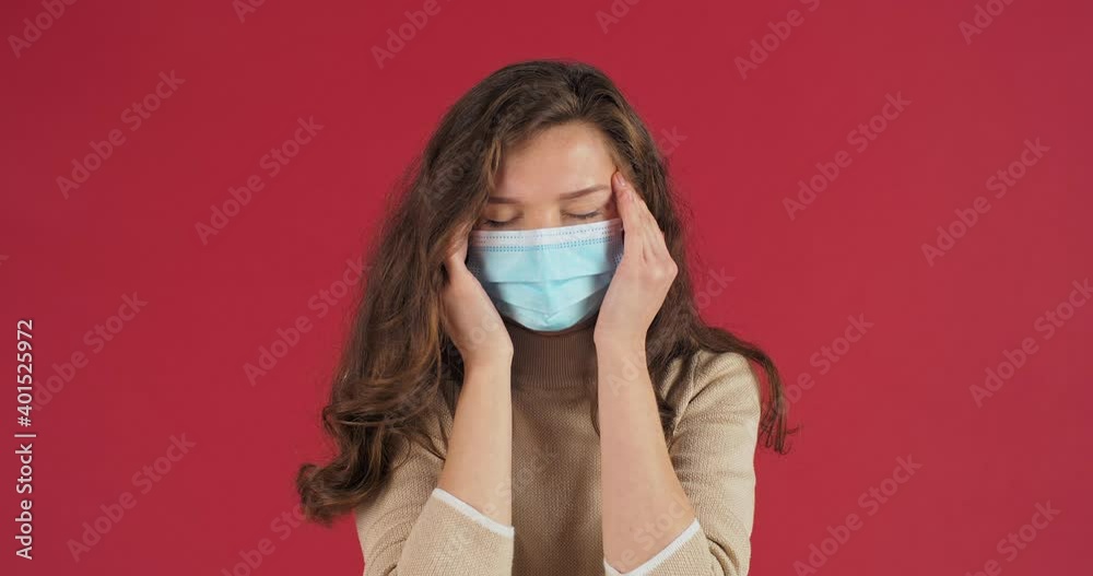 Portrait of female sad face portrait in studio on red background young woman in protective medical mask feels headache suffers from migraine high temperature holds her head with hands disease concept Portrait of female sad face portrait in studio on red background young woman in protective medical mask feels headache suffers from migraine high temperature holds her head with hands disease concept
