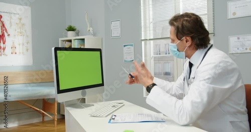 Wallpaper Mural Portrait of Caucasian male general practitioner in mask sitting at table in clinic room, speaking though webcam on computer looking at green screen. Monitor with chroma key. Doctor online consultation Torontodigital.ca
