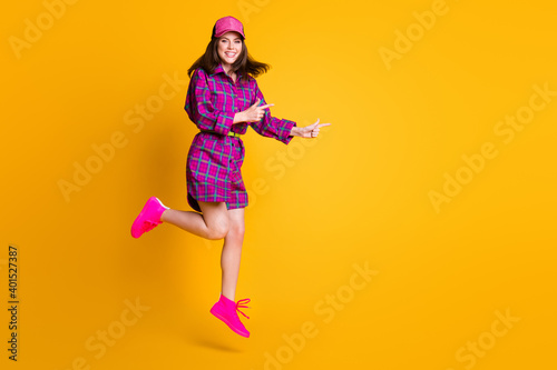 Full body portrait of pretty girl jump finger point empty space headwear plaid outfit isolated on yellow color background