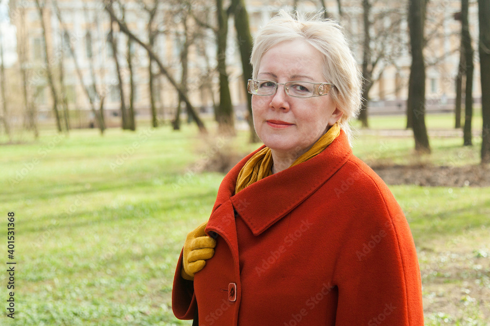 Mature blonde shorthaired woman in red coat, golden glasses and yelllow glowes in spring park Mature blonde shorthaired woman in red coat, golden glasses and yelllow glowes in spring park