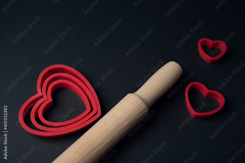 Red heart-shaped baking tins for cookies and wooden rolling pin on the dark (black) background. Flat lay, top view, space for text. Valentine's day concept. Red heart-shaped baking tins for cookies and wooden rolling pin on the dark (black) background. Flat lay, top view, space for text. Valentine's day concept.