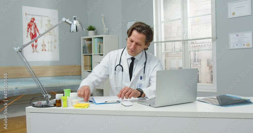 Portrait of busy serious middle-aged Caucasian male healthcare specialist sitting in hospital room, working and writing down information looking at laptop screen waiting for a patient Medicine concept Portrait of busy serious middle-aged Caucasian male healthcare specialist sitting in hospital room, working and writing down information looking at laptop screen waiting for a patient Medicine concept