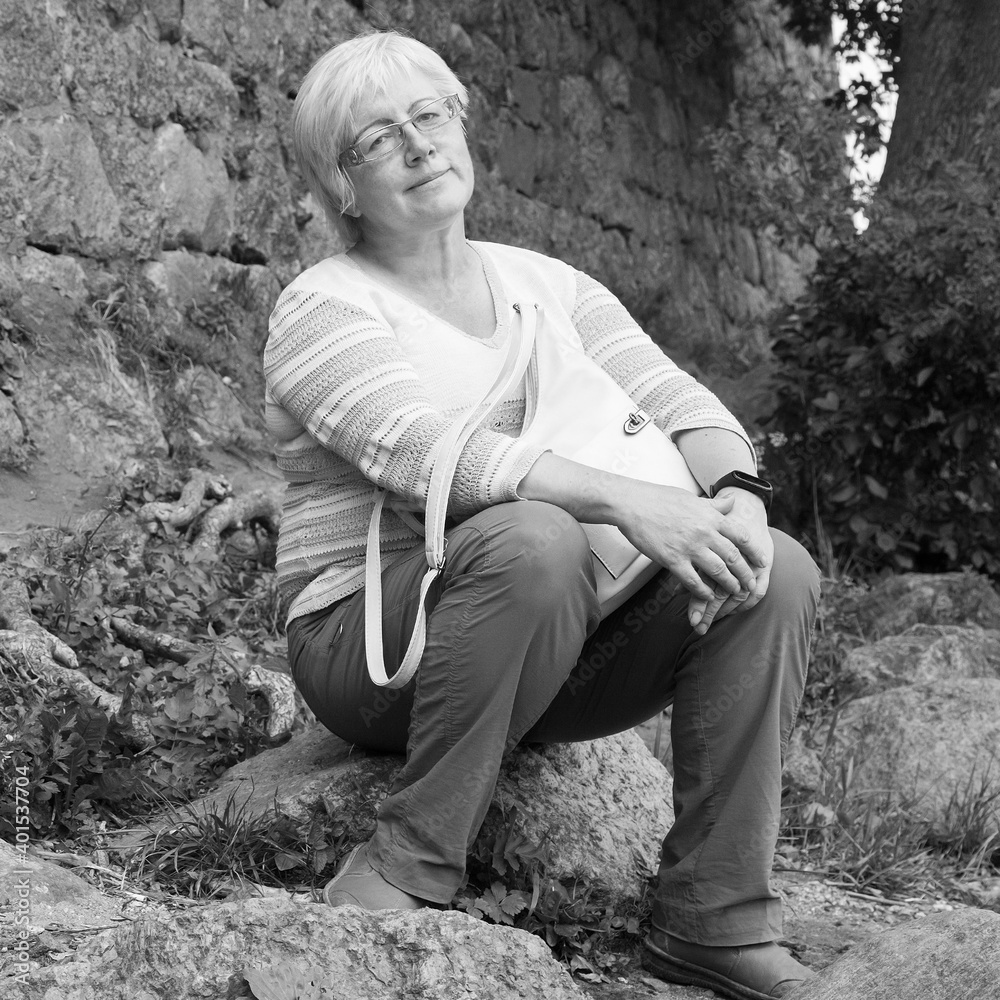 Mature woman in glasses with short blond hair in linen clothes..  sitting on a granite stone near castle wall