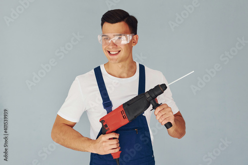 In protective eyewear and with drill in hands. Male worker in blue uniform standing inside of studio against white background
