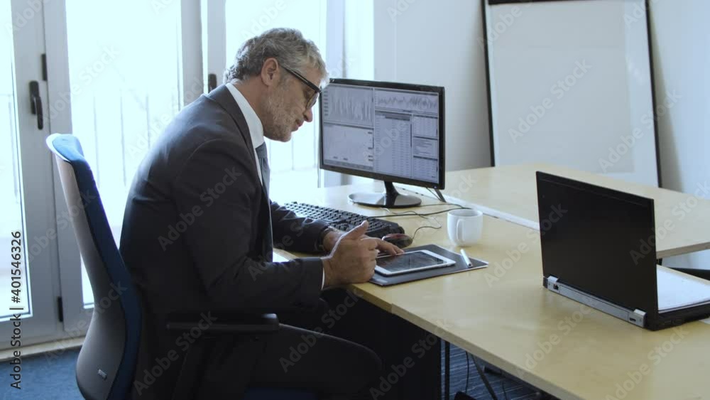 Focused grey haired experienced investor sitting and working at monitor with financial charts, analyzing data and using tablet. Dolly shot. Business and stock market concept Focused grey haired experienced investor sitting and working at monitor with financial charts, analyzing data and using tablet. Dolly shot. Business and stock market concept