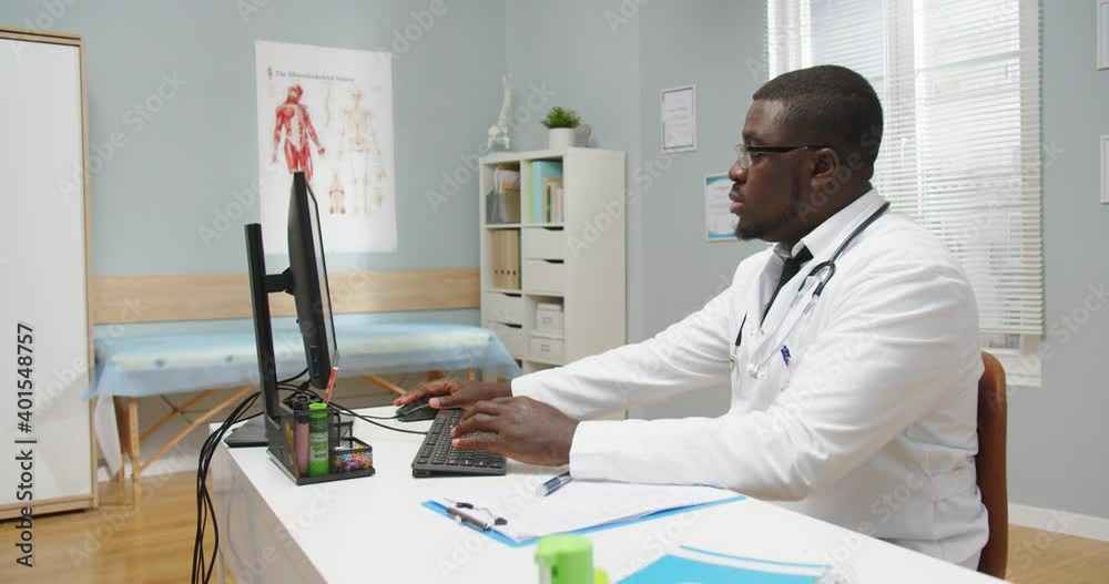 Side view. Portrait of serious African American male doctor in white coat sitting at desk in cabinet browsing and tapping on computer, writing down information in clinical history alone Health concept Side view. Portrait of serious African American male doctor in white coat sitting at desk in cabinet browsing and tapping on computer, writing down information in clinical history alone Health concept