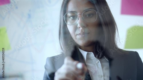 Focused young Latin business woman drawing charts and writing on glass board with sticky notes. Front view. Business plan concept