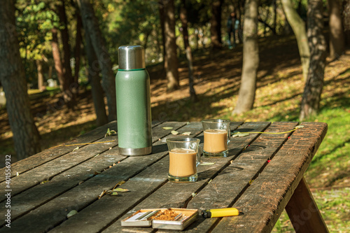 a coffee break in forest park, green steel thermos and two glass cups of coffee on wooden table. 