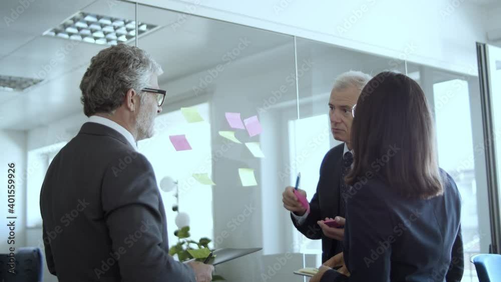 Company leaders writing and sticking paper notes on glass board, discussing project strategy. Low angle. Business plan concept Company leaders writing and sticking paper notes on glass board, discussing project strategy. Low angle. Business plan concept