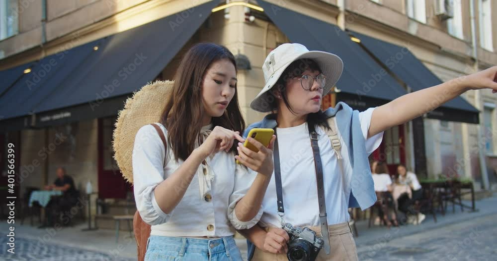 Portrait of young Asian beautiful female standing on street and searching for direction while tapping on cellphone. Woman in hat and glasses pointing at something while looking away. Travel concept
