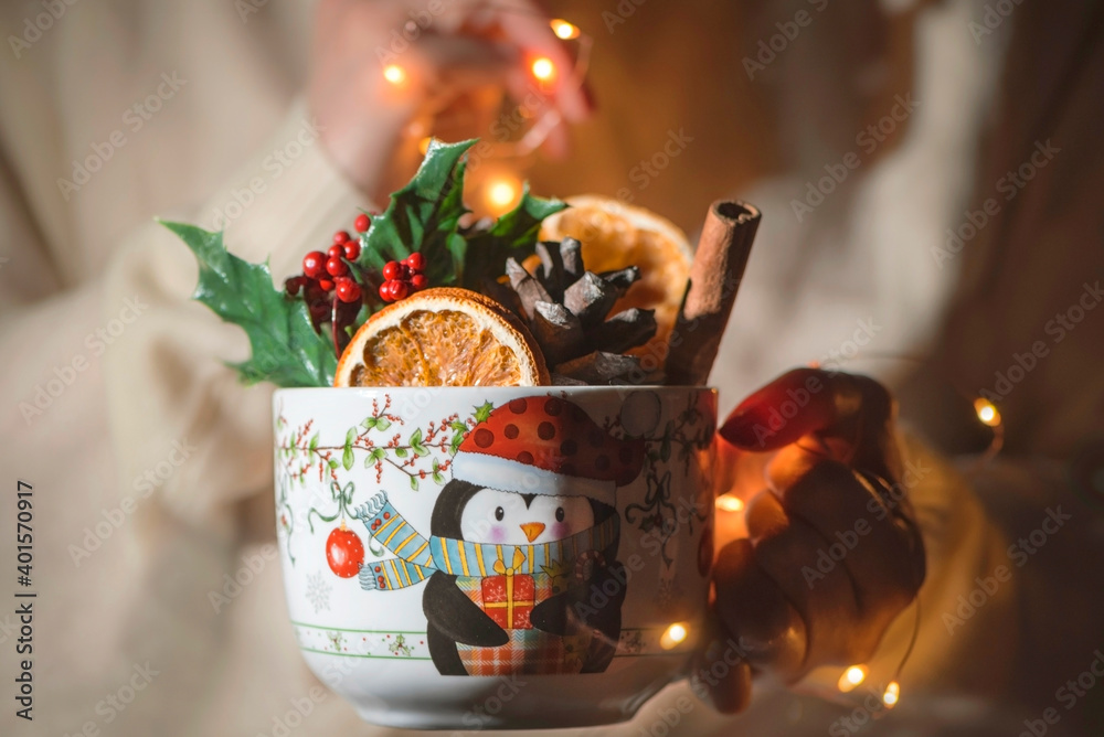 A young woman holds large mug with full of Christmas flower, oranges, cinnamon sticks and pine cone with lights , close up, still life photography