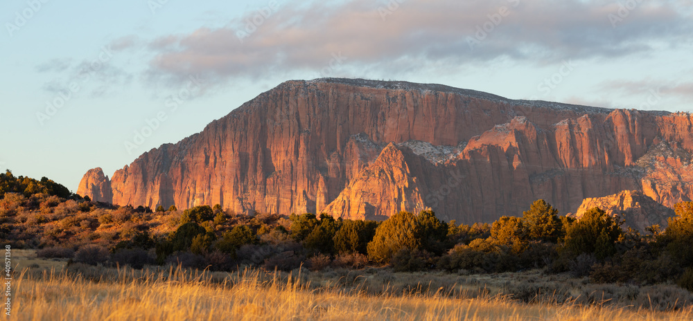 Kolob fingers mountains are lit by warm evening light late on a winter day seen from a remote dirt road with grassy fields and juniper trees in the foreground.