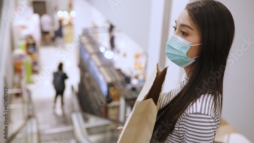 Young Asian woman in medical mask grocery walk toward camera shot, at supermarket during covid-19 coronavirus pandemic. Young lady stocks up food and toilet paper.