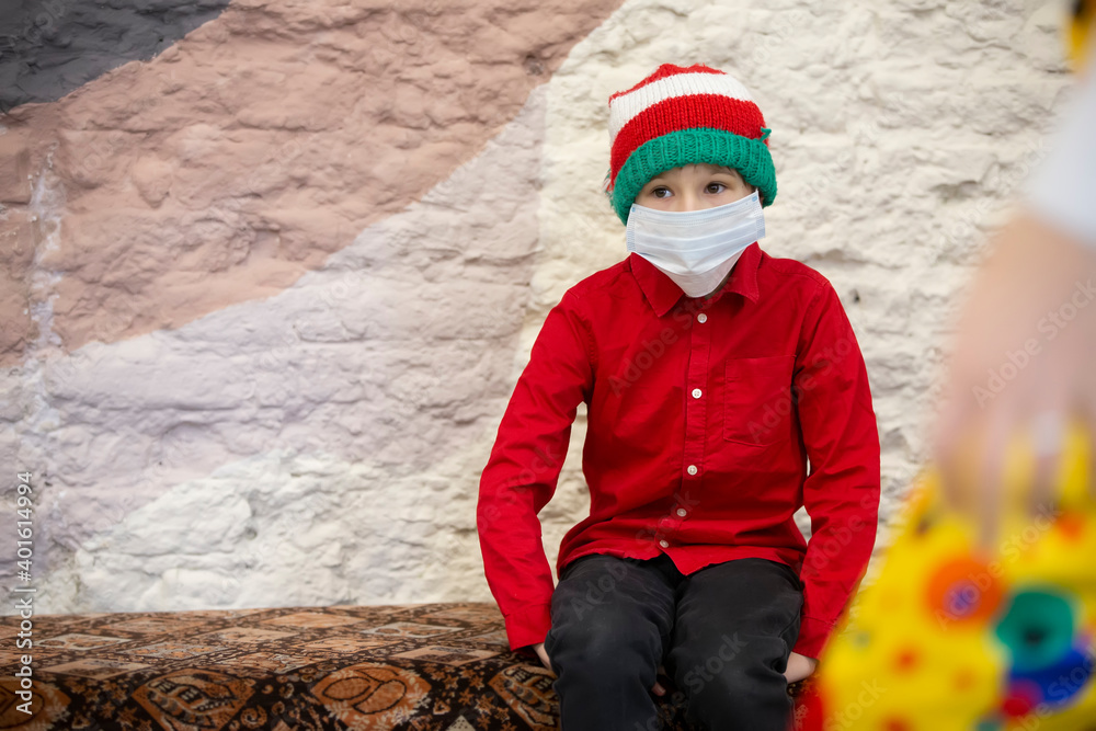 A child in a medical mask wearing a Santa Claus hat due to the coronavirus pandemic. Sad little boy wearing a Christmas hat during the coronavirus epidemic. A child in a medical mask wearing a Santa Claus hat due to the coronavirus pandemic. Sad little boy wearing a Christmas hat during the coronavirus epidemic.