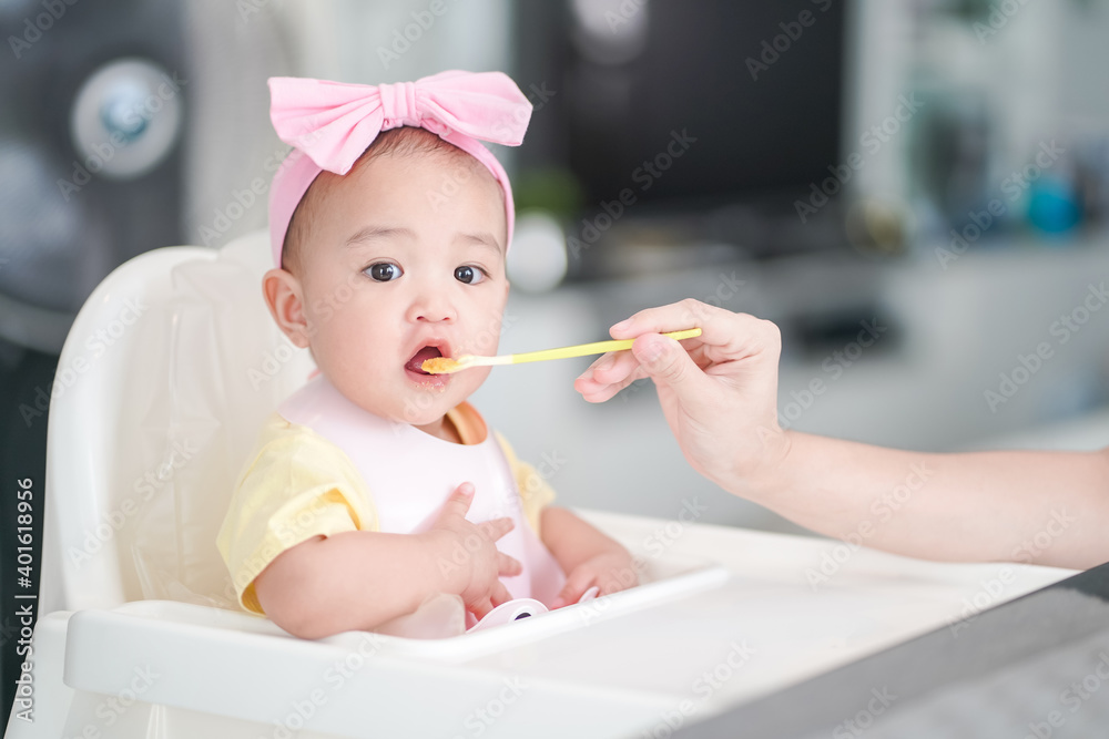 Asian cute baby girl in a pink bib and hair bow sitting on a white high chair. Her mother giving healthy food by spoon in the morning at home Asian cute baby girl in a pink bib and hair bow sitting on a white high chair. Her mother giving healthy food by spoon in the morning at home