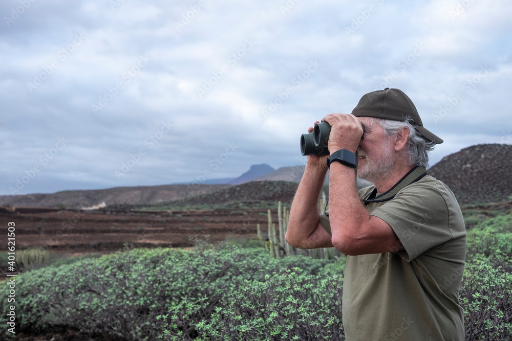 A white-haired smiling senior man with cap looking away the mountain landscape with binoculars enjoying freedom in nature A white-haired smiling senior man with cap looking away the mountain landscape with binoculars enjoying freedom in nature