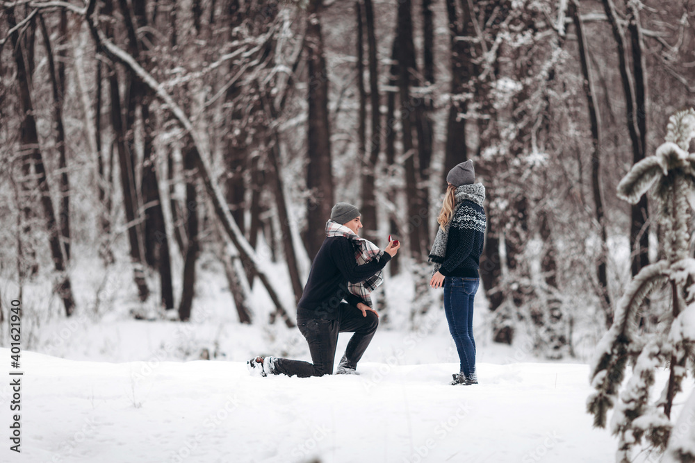 A guy kneeling down puts a wedding ring on a girl's hand, making a proposal to marry in a snowy forest in winter