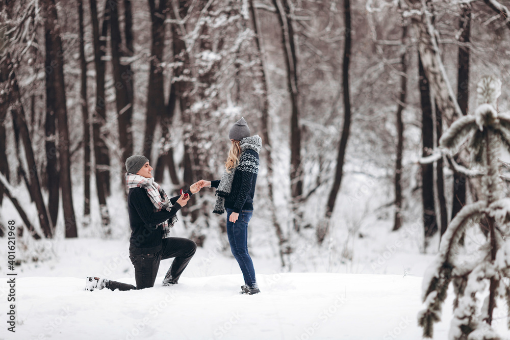 Naklejka premium Kneeling guy puts an engagement ring on a girl's hand, making a proposal to get married in winter in a snowy forest