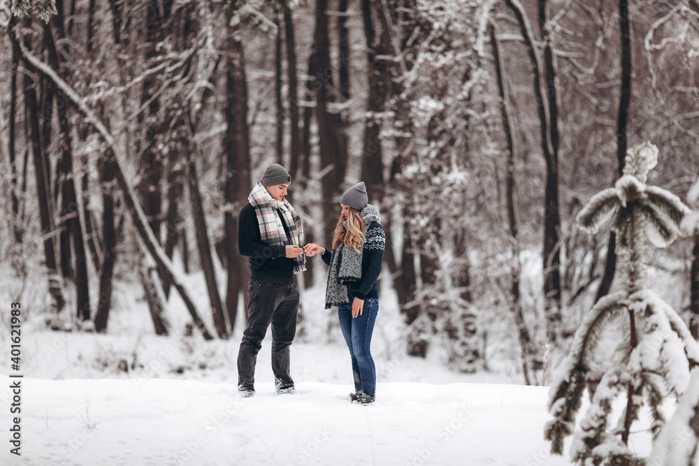 A guy kneeling down puts a wedding ring on a girl's hand, making a proposal to marry in a snowy forest in winter