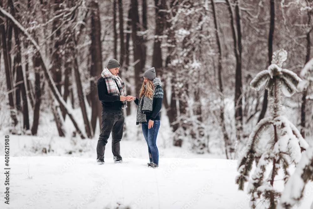 A guy kneeling down puts a wedding ring on a girl's hand, making a proposal to marry in a snowy forest in winter