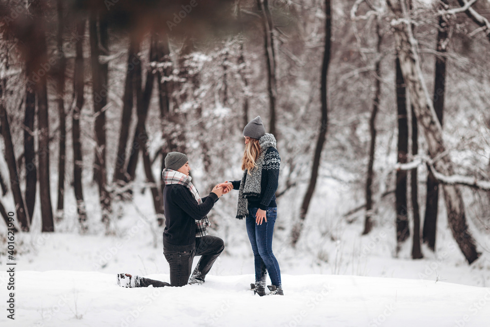 Kneeling guy puts an engagement ring on a girl's hand, making a proposal to get married in winter in a snowy forest