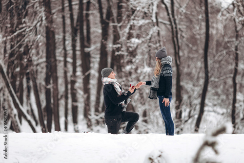 A guy kneeling down puts a wedding ring on a girl's hand, making a proposal to marry in a snowy forest in winter
