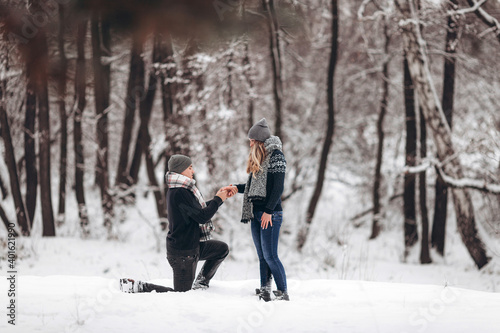 Kneeling guy puts an engagement ring on a girl's hand, making a proposal to get married in winter in a snowy forest © Aleksandr