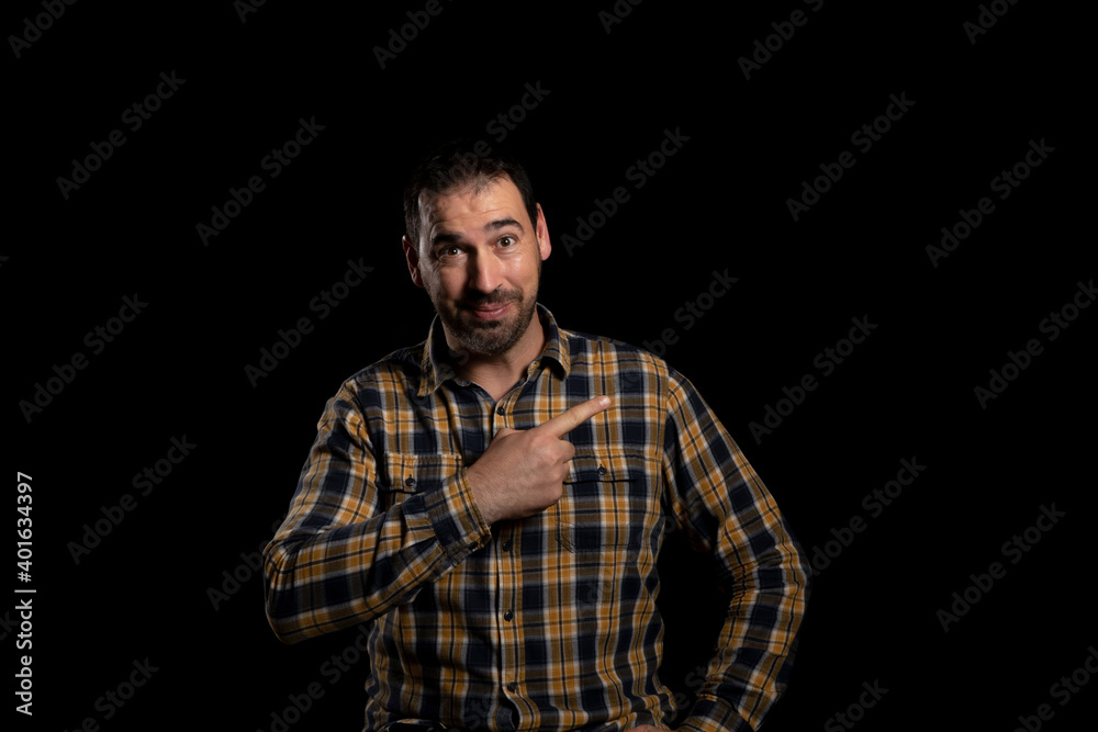 Young handsome man, wearing a stylish yellow shirt on an isolated black studio background, surprised, smiling at the camera and pointing a finger