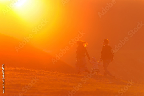 Back view of anonymous parents holding hands of child and running on hill towards bright sundown sun in weekend in countryside