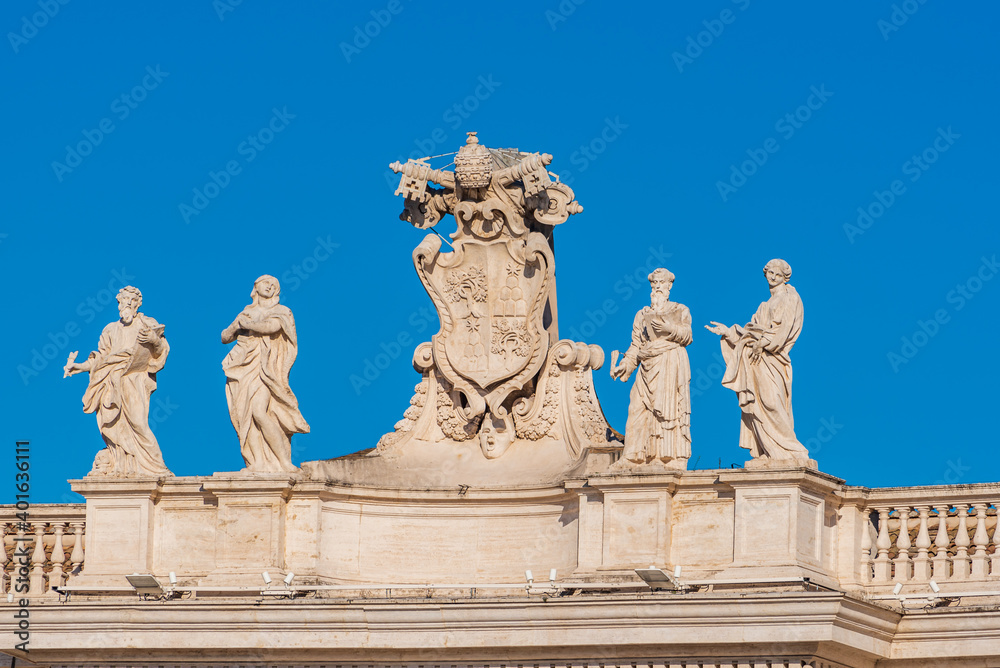 Marble statues standing  next to Vatican City coat of arms on the roof top of building 