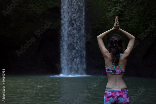 Hands raising up in namaste mudra. Woman meditating  practicing yoga and pranayama with namaste mudra near waterfall. Yoga outdoor concept. Tibumana waterfall  Bali. Copy space. View from back.