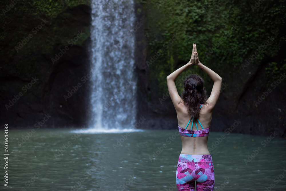 Hands raising up in namaste mudra. Woman meditating, practicing yoga and pranayama with namaste mudra near waterfall. Yoga outdoor concept. Tibumana waterfall, Bali. Copy space. View from back.