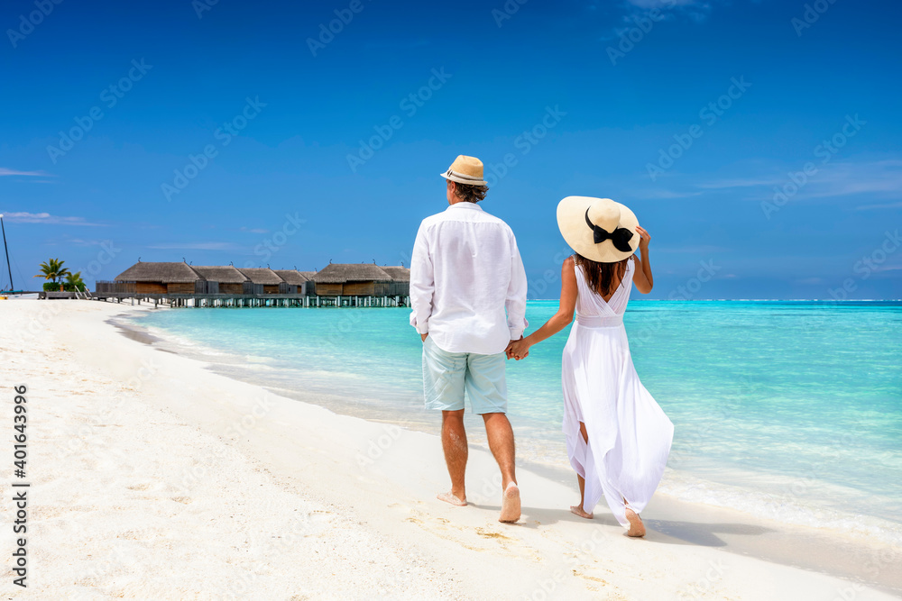 Happy couple in white clothing and with hats walks down a tropical beach with turquoise sea in the Maldives islands