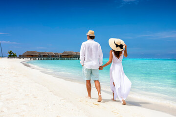 Happy couple in white clothing and with hats walks down a tropical beach with turquoise sea in the Maldives islands