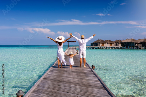 A happy family in white summer clothing on vacation walks along a wooden pier over tropical, turquoise ocean in the Maldives, Indian Ocean